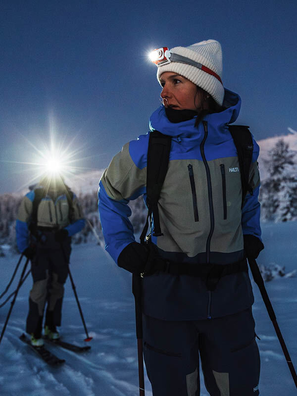 Two people in Halti Rizle Unisex 3L DX Ski Jackets cross-country ski a snowy, mountainous landscape at dusk.
