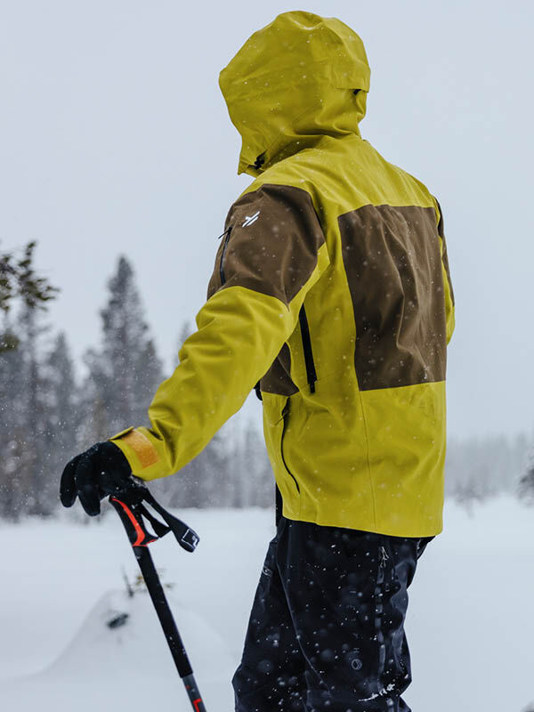 A person wearing a lime green and brown Halti Rizle Unisex 3L DX Ski Jacket holds a ski pole in a snowy landscape.