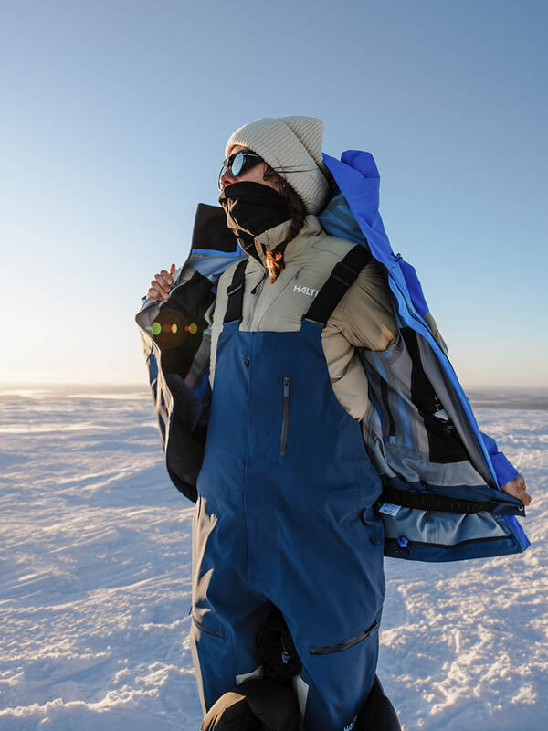 A person in a blue Rizle Unisex 3L DX Ski Jacket over a tan jacket and blue ski bibs stands in a snowy landscape.