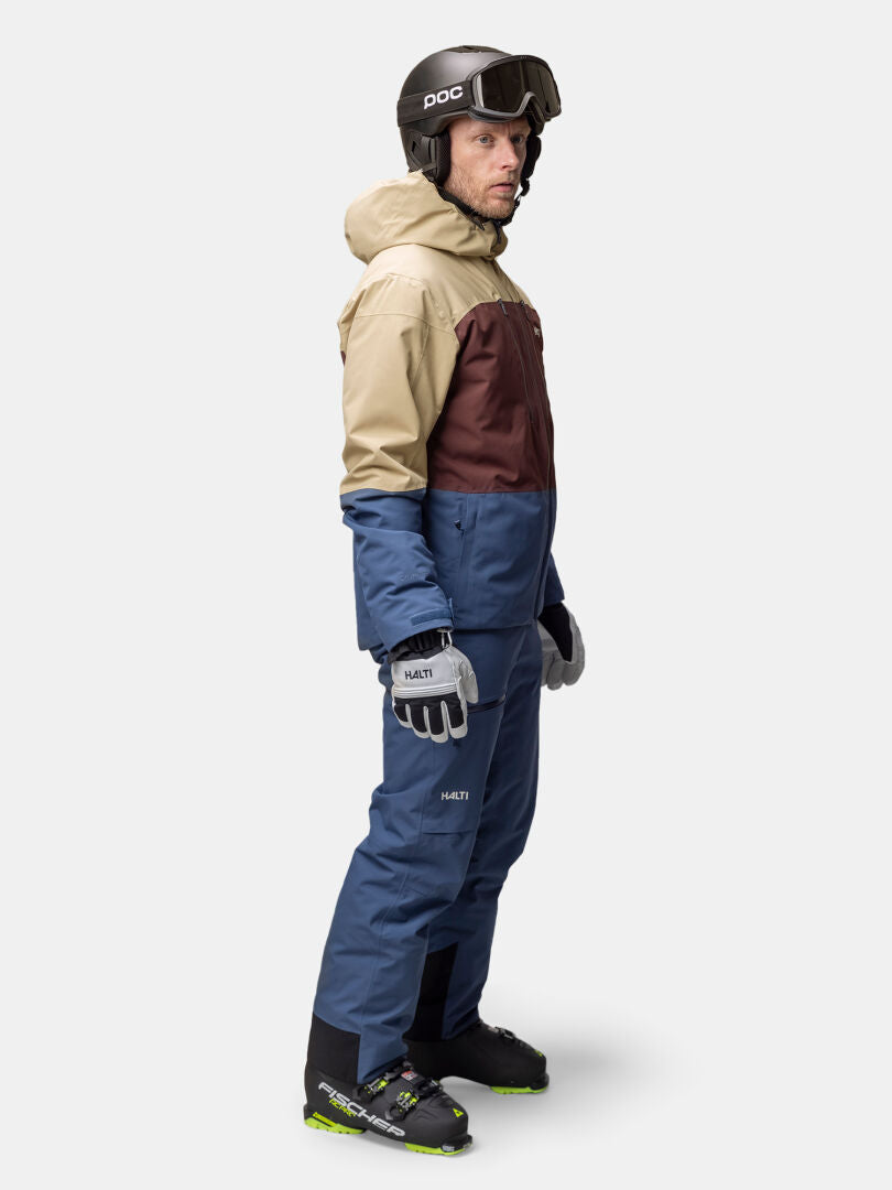 A man in a helmet and goggles wears the Halti Melvik Dx Ski Jacket Men's, blue ski pants, gloves, and ski boots.