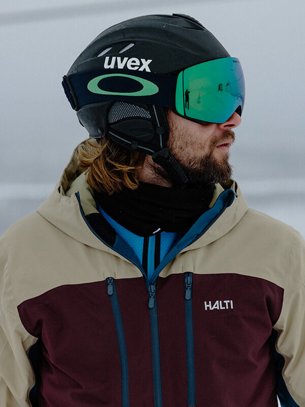 A man in the Halti Melvik Dx Ski Jacket and green goggles faces sideways in a snowy outdoor setting.