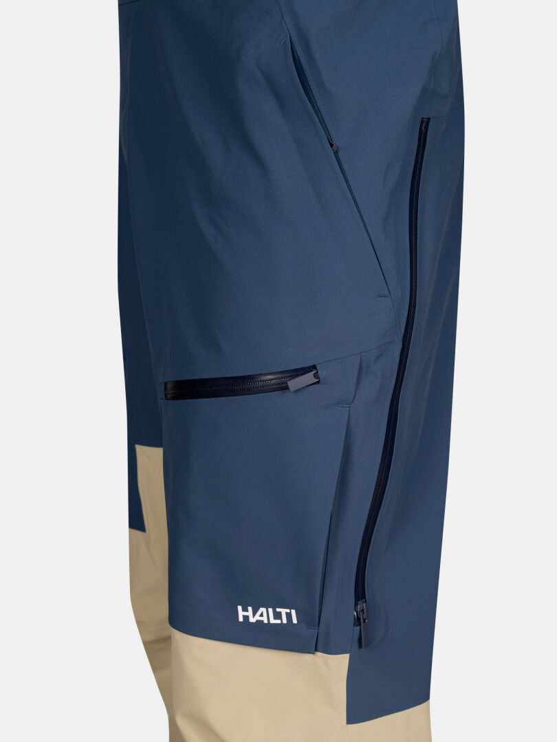 Close-up of blue and beige Rizle 3L Dx Ski Pants Unisex by Halti, with zippered pockets and branding near the knee.