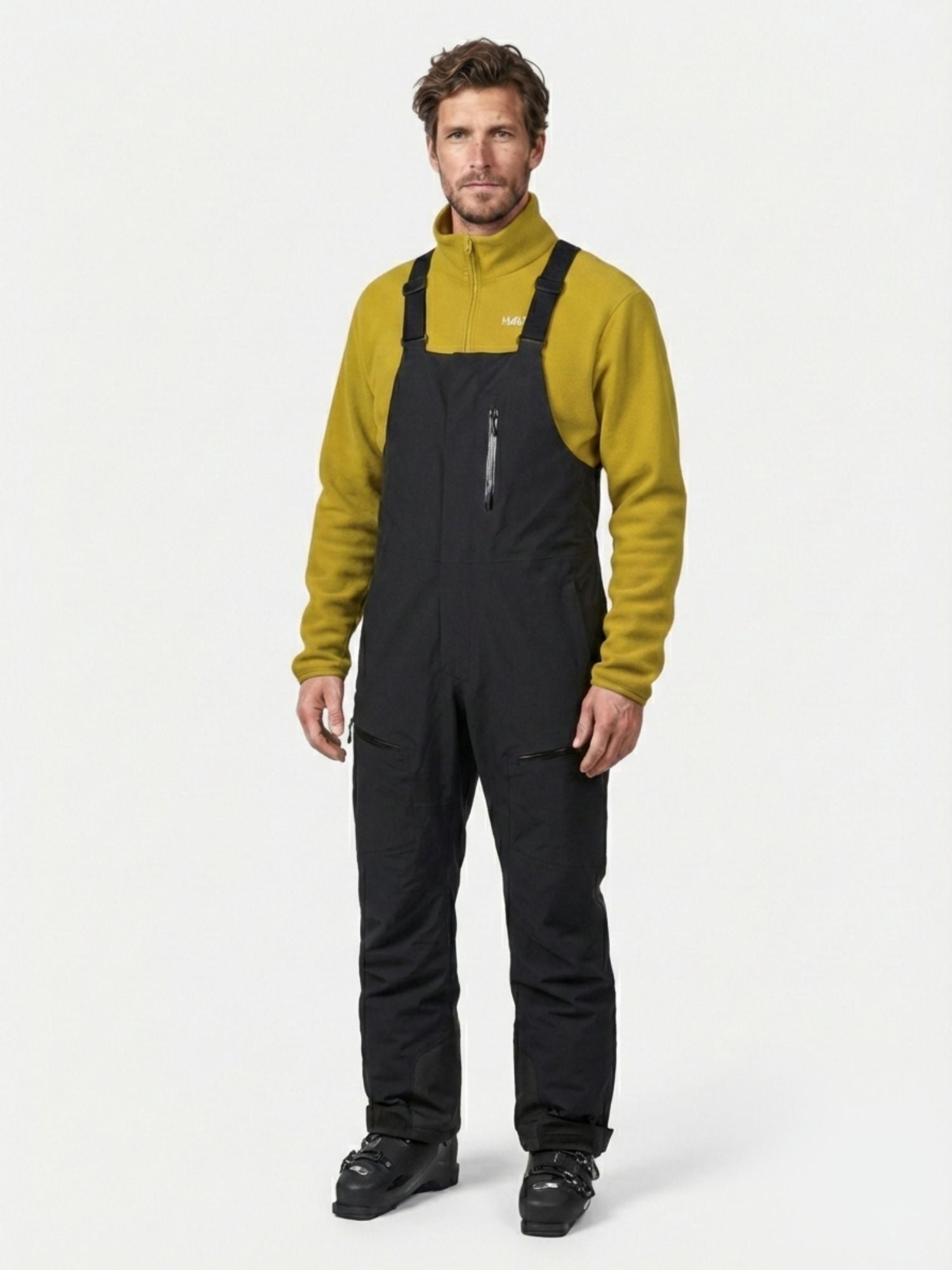 A man wears Halti Rizle 3L Dx Unisex Ski Pants, a yellow fleece, and black ski boots against a plain background.