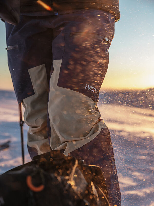 Close-up of a person wearing Halti Rizle 3L Dx Ski Pants Unisex on snowy terrain at sunset, snow particles in the air.