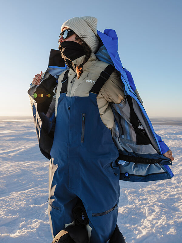 In Halti Rizle 3L Dx Ski Pants, person in winter gear opens blue jacket on snowy landscape under low sun.