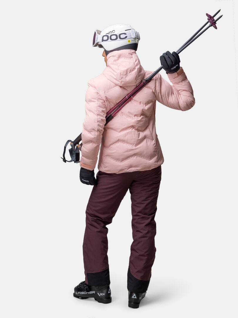 A woman wears the Halti Nordic Arcty II Ski Jacket, holding ski poles over her shoulder against a white background.