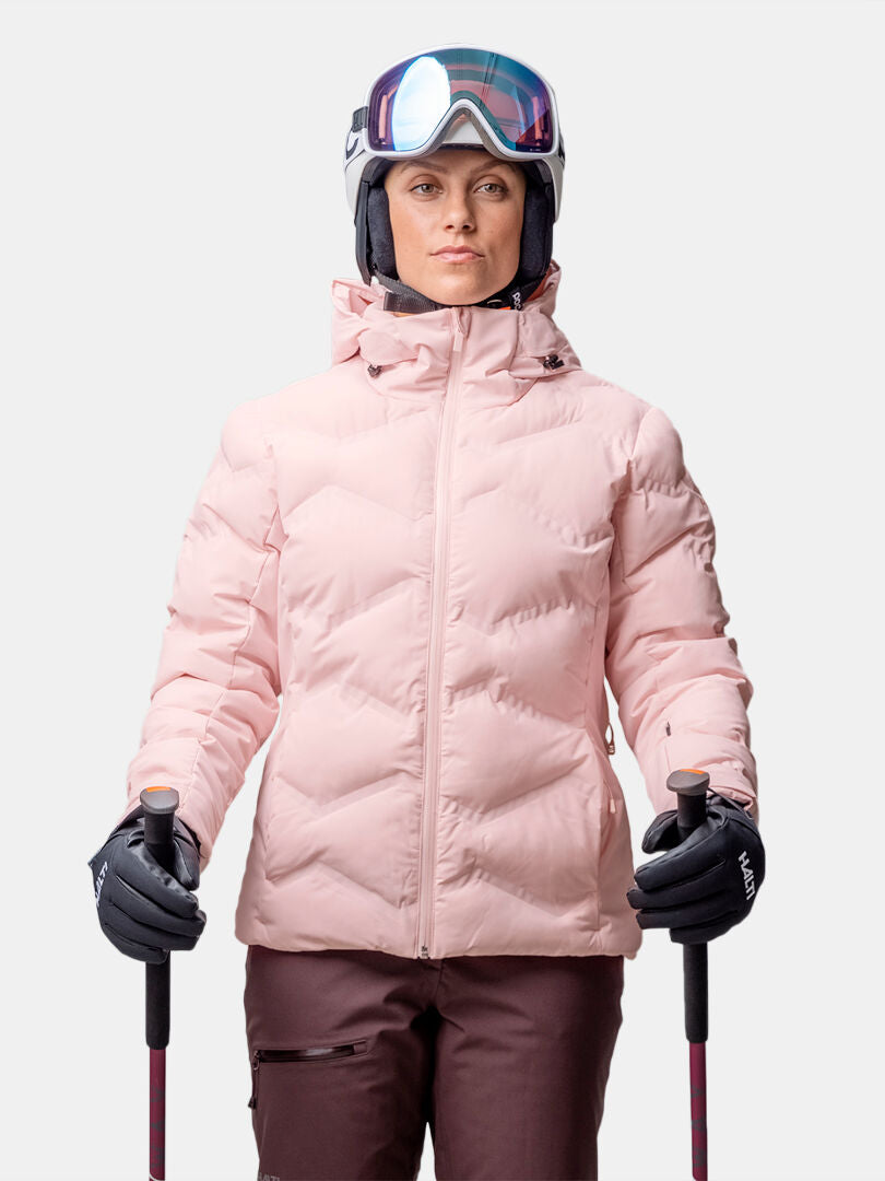 A person in a Halti Nordic Arcty II Women's Ski Jacket, brown pants, helmet, goggles, and gloves holds ski poles.