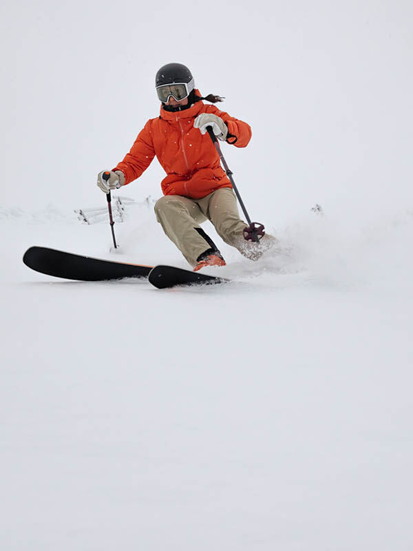 Woman in Halti Nordic Arcty II Ski Jacket skiing downhill, helmet on, spraying snow on a snowy slope.