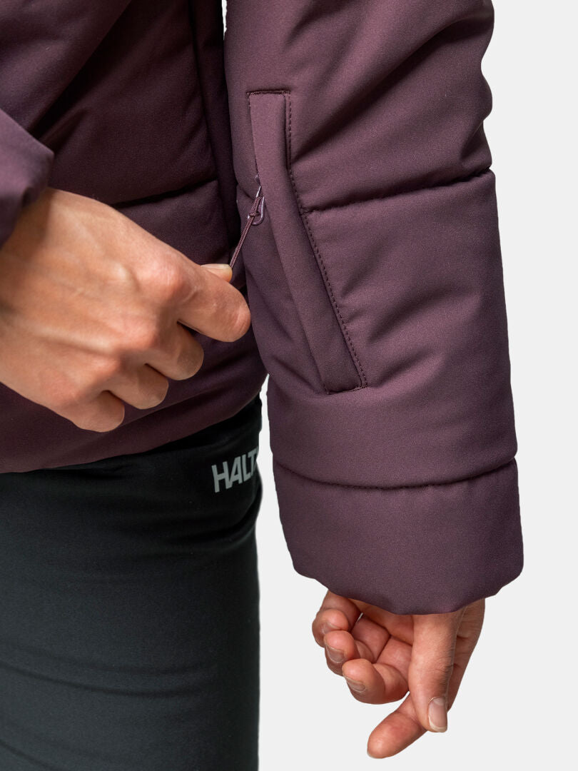 A woman unzips a sleeve pocket on the purple Halti Ascale Puffer Ski Jacket, paired with black pants.