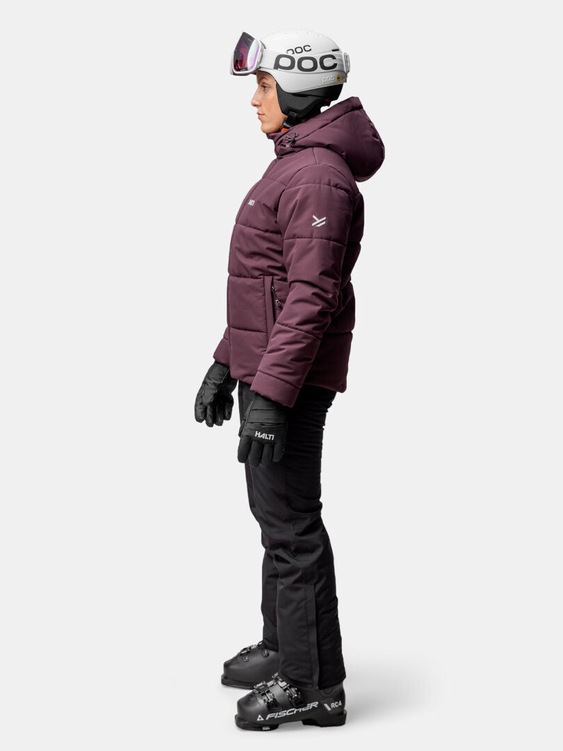 Woman in full ski gear, wearing Halti Ascale Puffer Ski Jacket, helmet, goggles, gloves, pants, and boots in profile view.