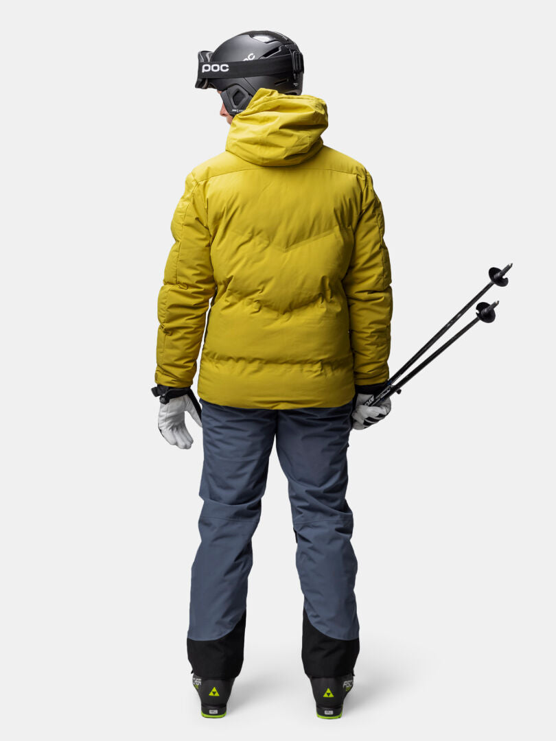 In a Halti Nordic Arcty II Ski Jacket, a man with gray pants and helmet stands holding ski poles, facing away.