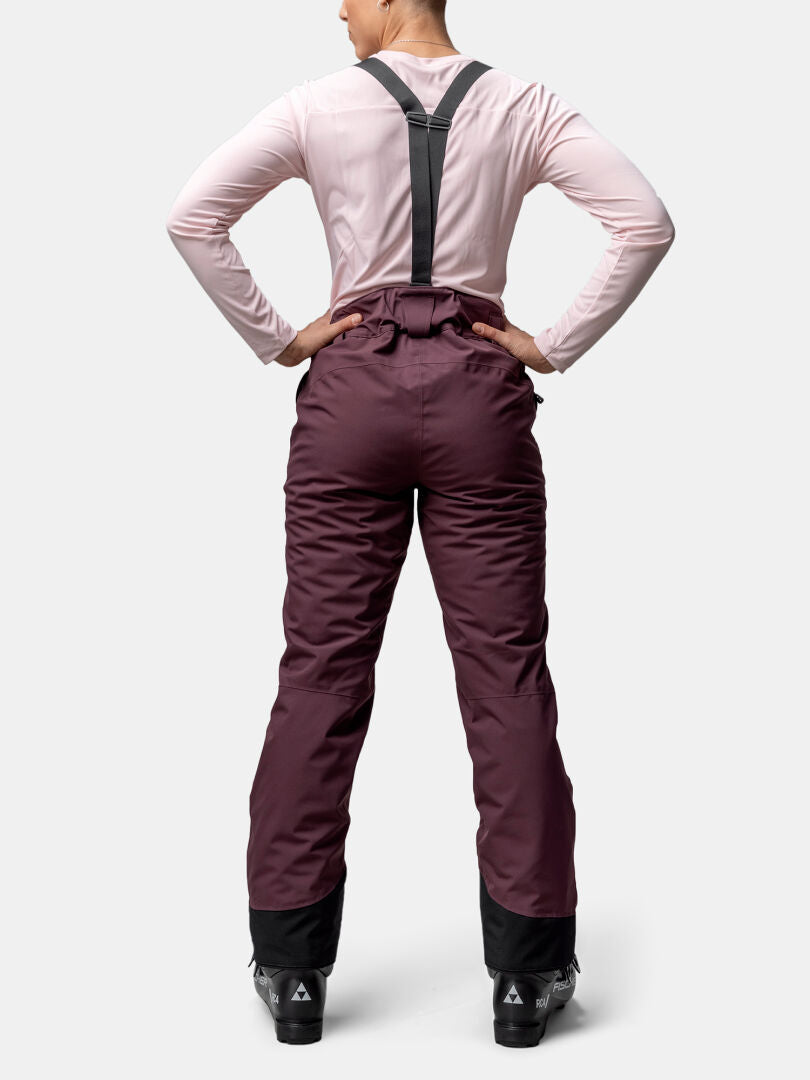 A person wears Halti Carvey II DX Women's maroon ski pants with black suspenders and boots, facing away hands on hips.
