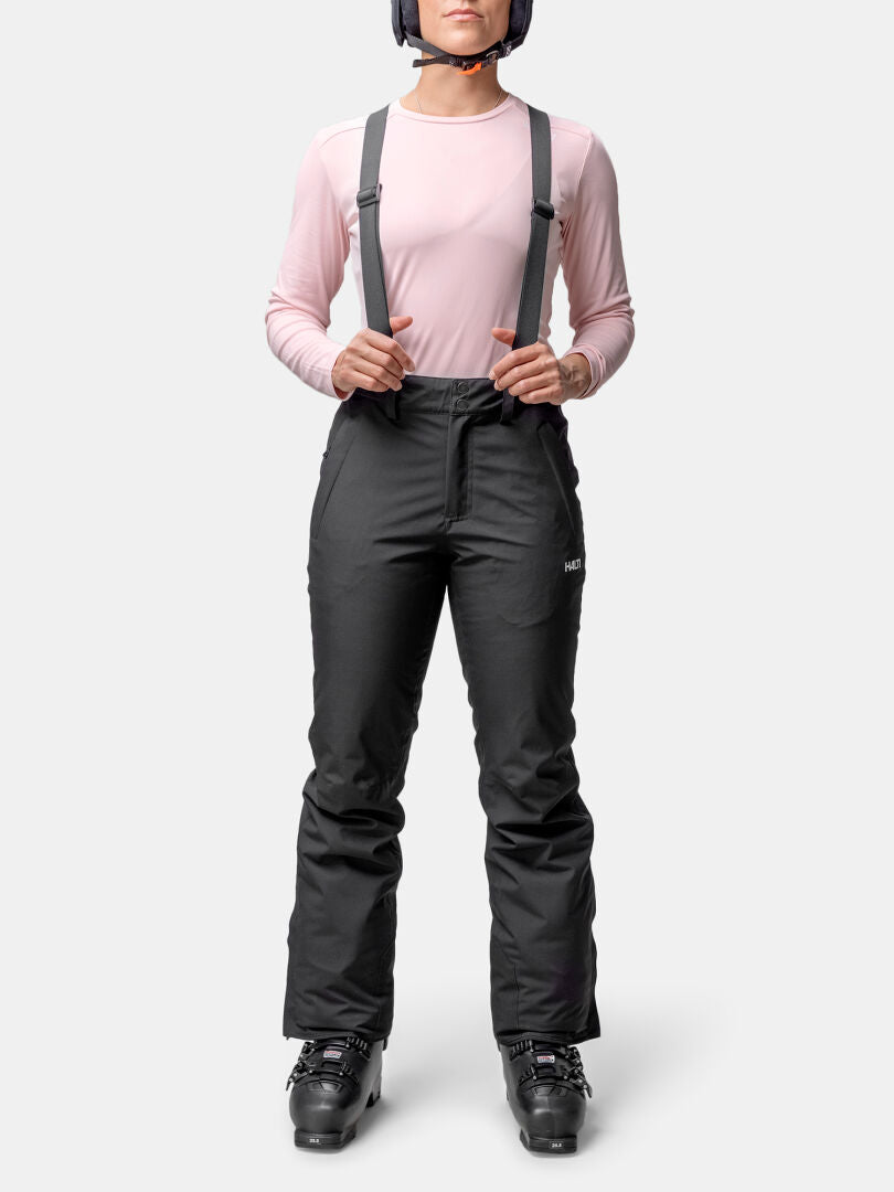 Woman in a pink long-sleeve shirt and Halti Glades II DX ski pants, black ski boots, and helmet, facing forward.