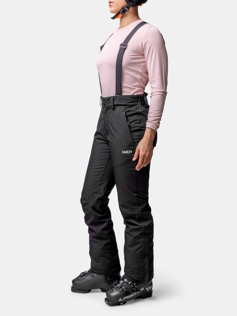 A person wears Halti Glades II DX Women's Ski Pants with suspenders, a pink long-sleeve shirt, ski boots, and a helmet.