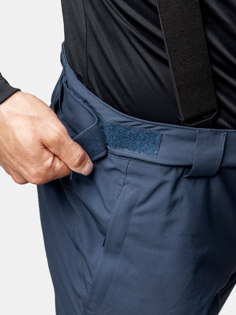 A person adjusts the Velcro waist of Halti Carvey II DX Men's navy ski pants while wearing a black shirt and suspenders.