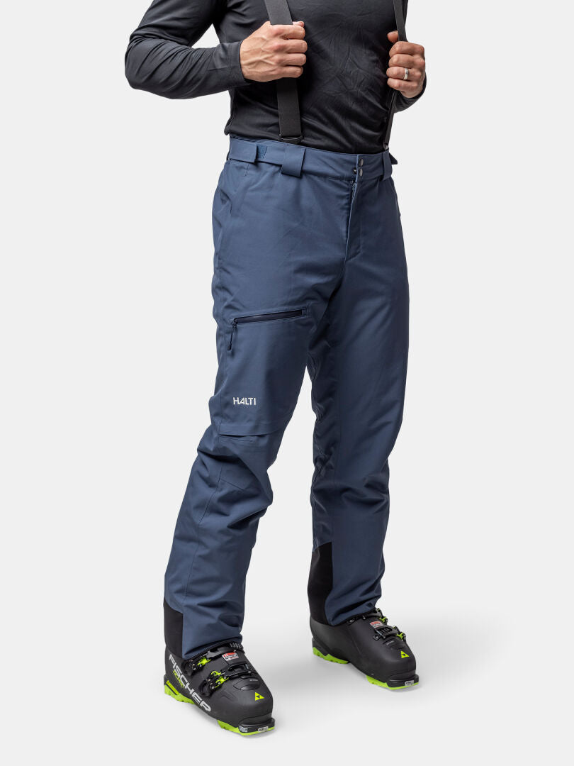 A man wears Halti Carvey II DX ski pants, black ski boots, and a long-sleeve shirt against a white background.