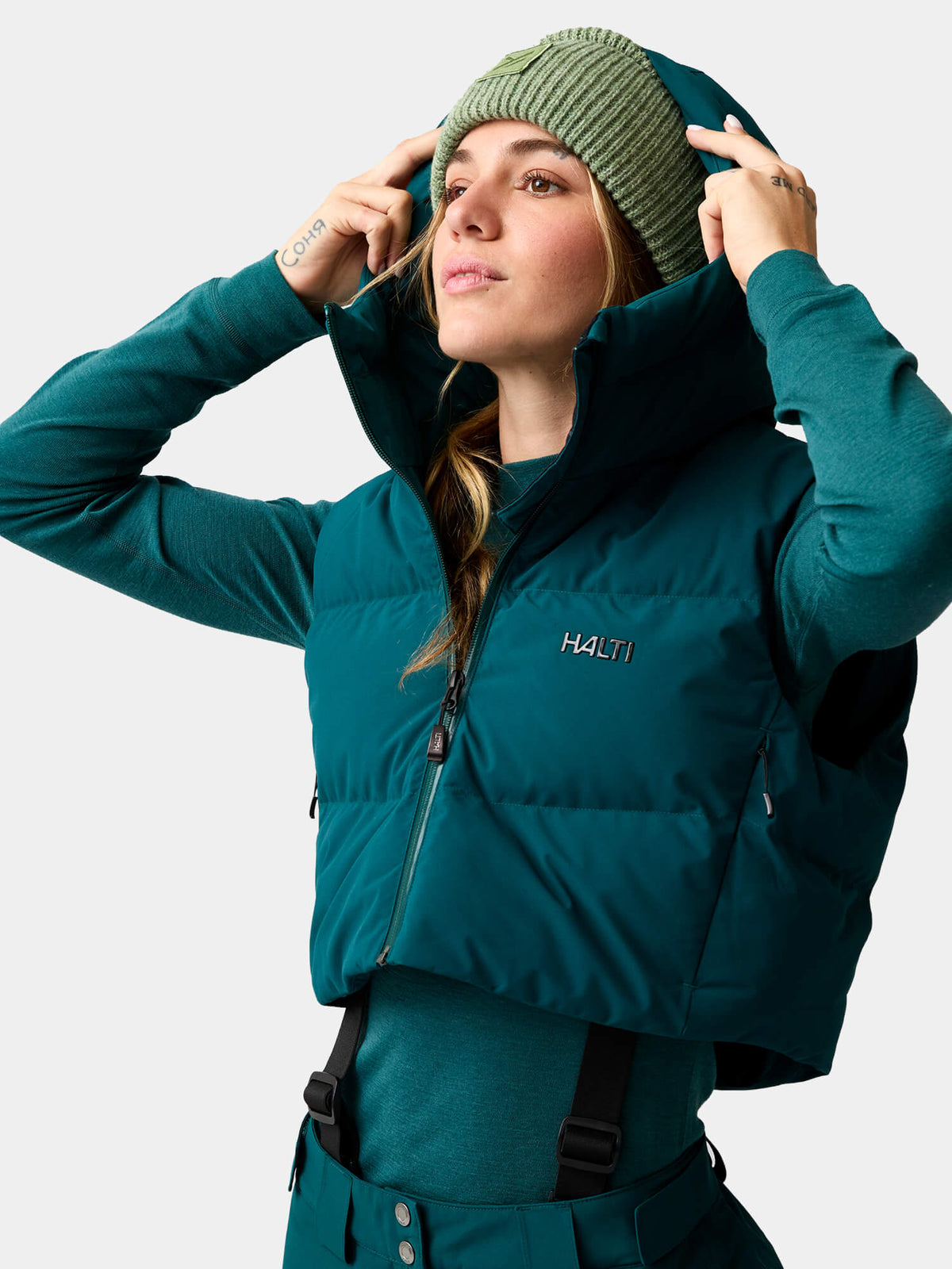 A woman in a dark teal Varion 2in1 DX Ski Jacket Women’s puffer vest, matching shirt, and green knit beanie.
