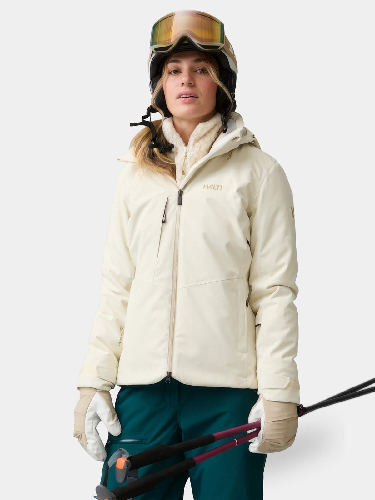A woman wears a waterproof off-white Halti Recier DX Ski Jacket, helmet, and goggles while holding ski poles.