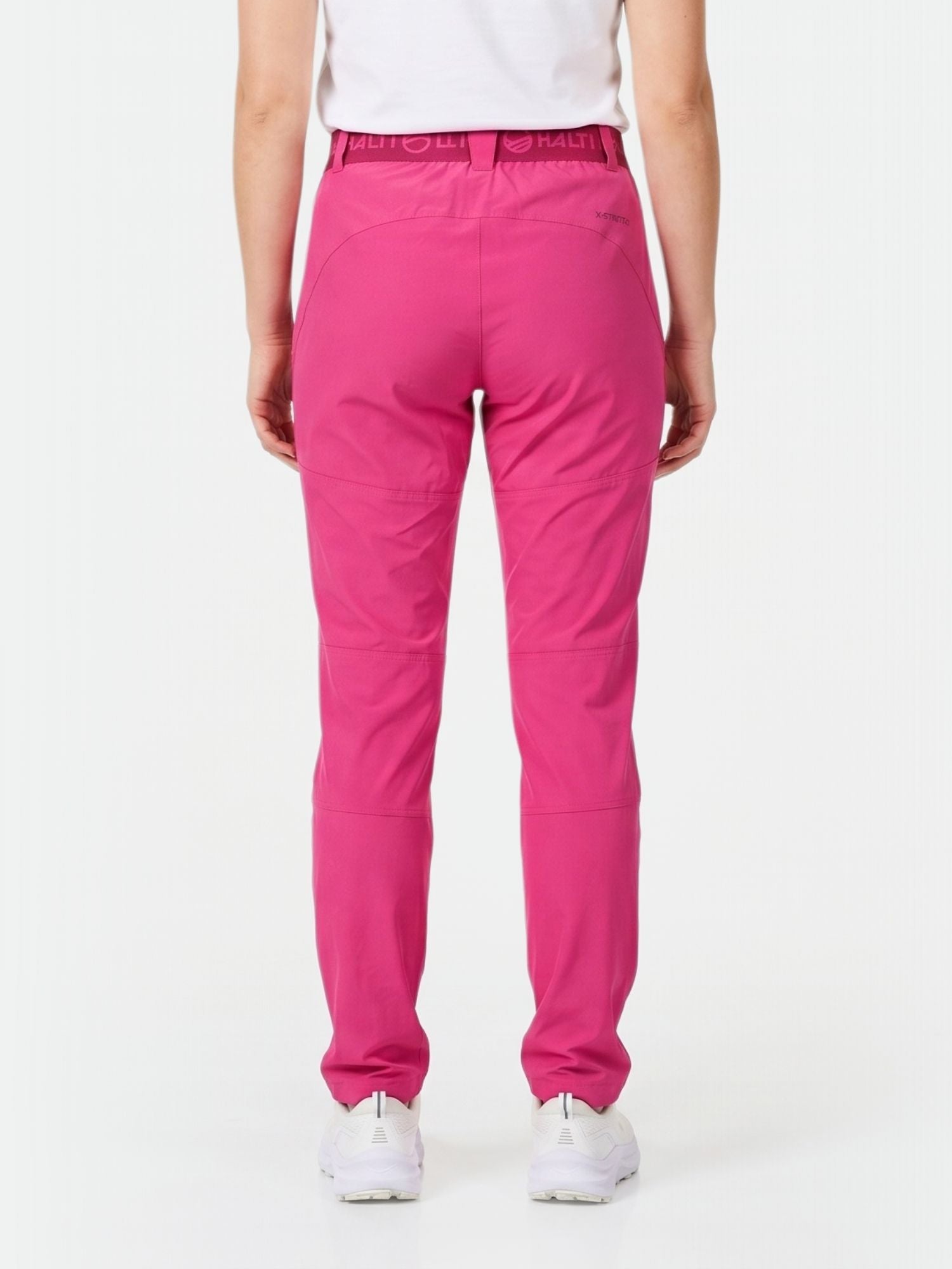 Model wears Halti Pallas Women's X-stretch Lite Pants in bright pink and white sneakers, seen from the back on white background.