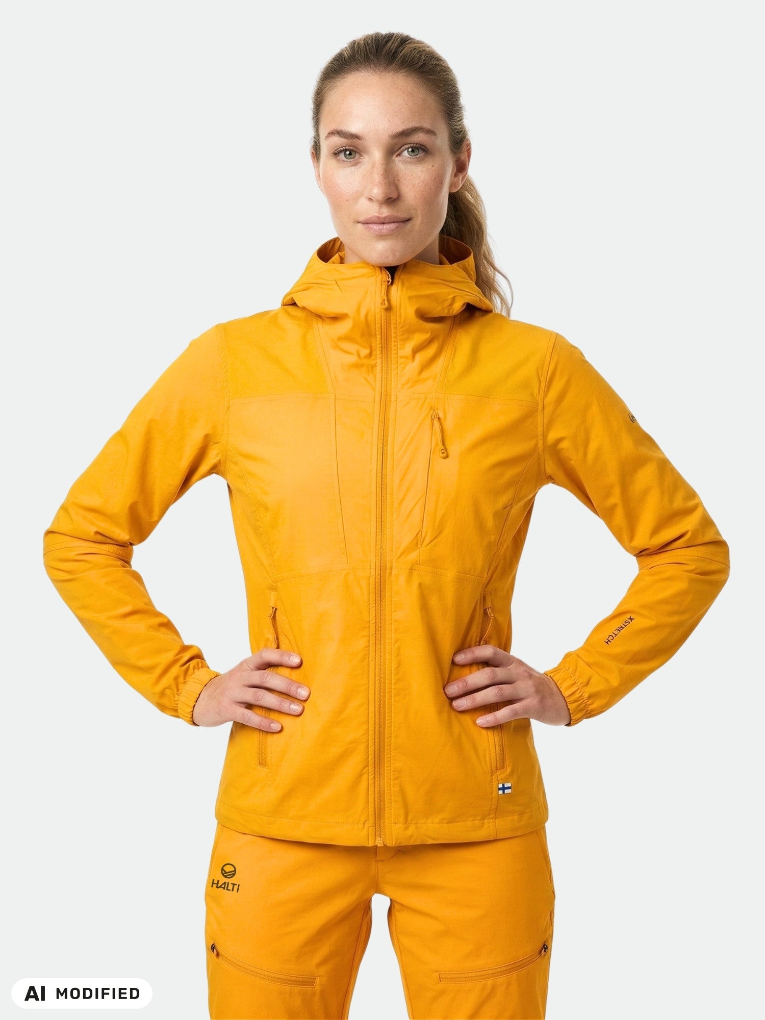 Woman wears a bright orange Halti Pallas II Women's X-stretch Jacket, hands on hips, against a white background.