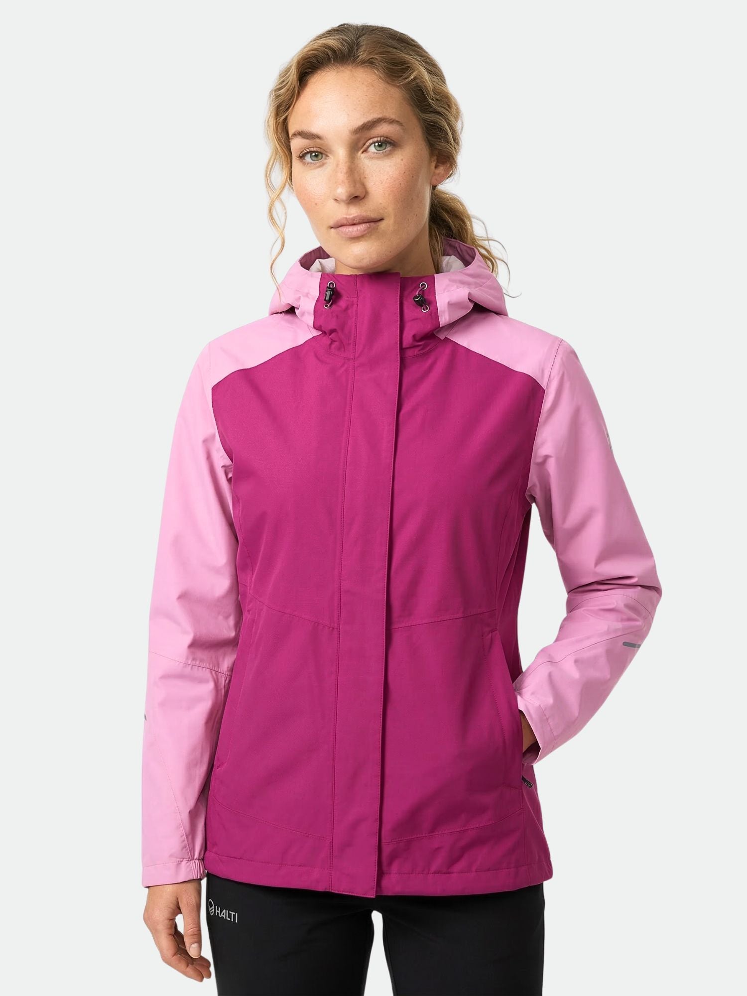 A woman wears the Halti Fort DX Shell Jacket Women's in magenta/pink with black pants, ideal for outdoor activities.