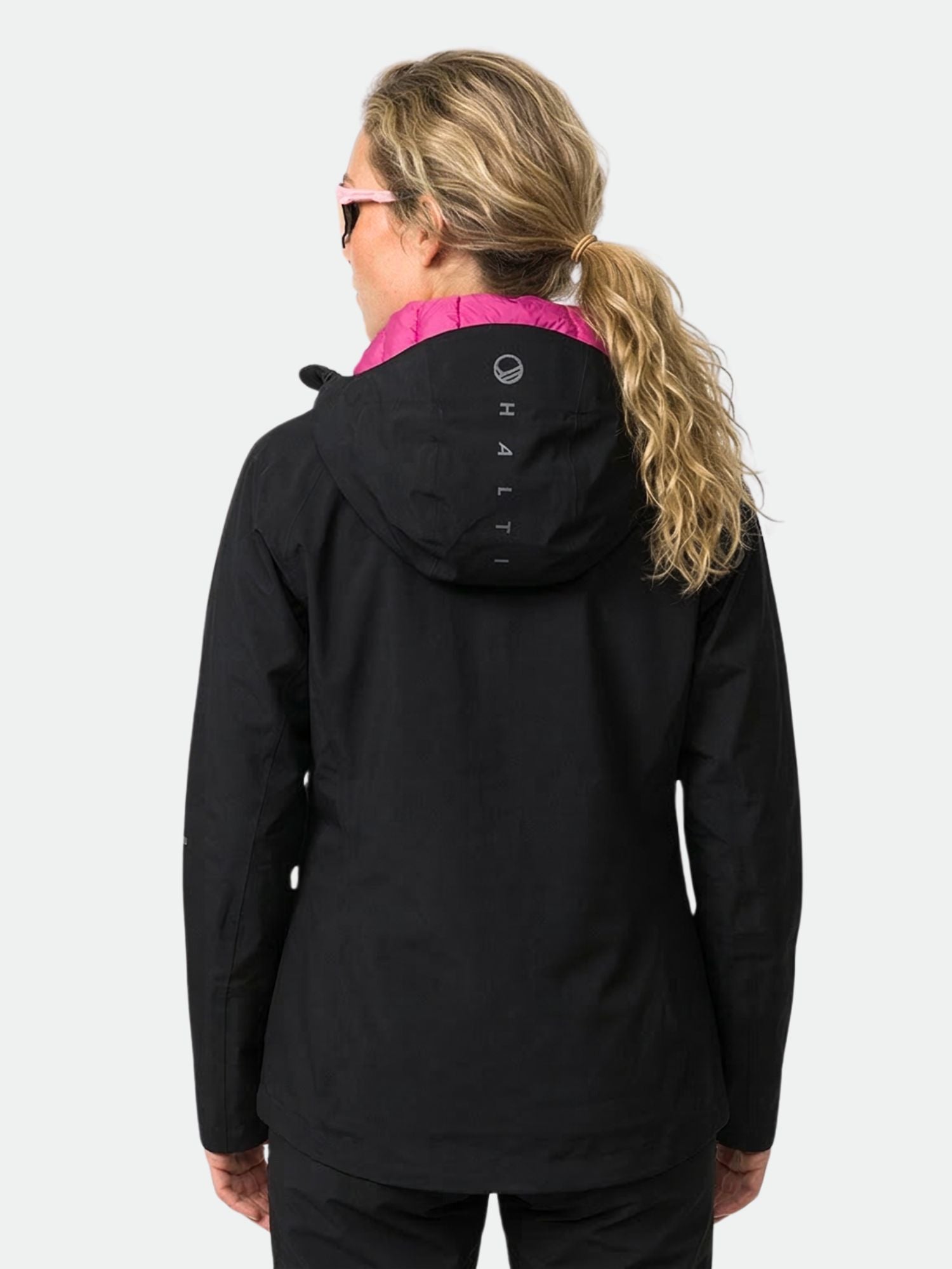 A woman with blond hair in a ponytail wears the Halti Kaarna DX 3L Shell Jacket Women's, black with pink collar, back view.