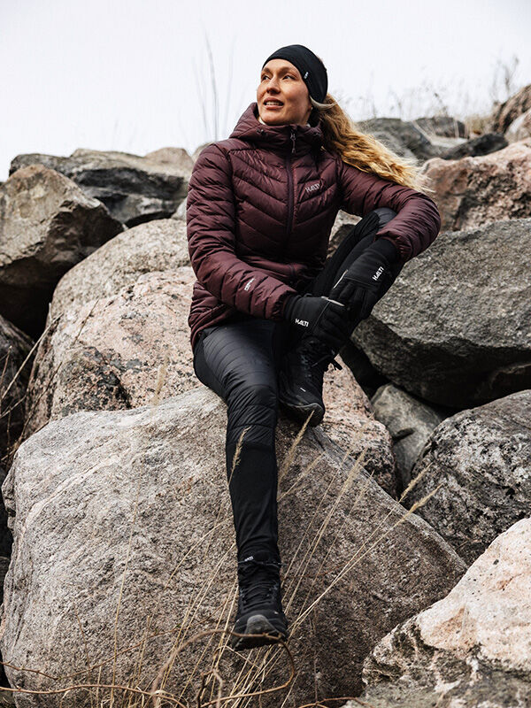 A woman in Halti Veidnes Insulation Pants sits on winter rocks, gazing sideways with a neutral expression.