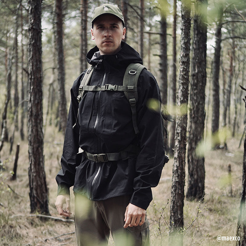 Wearing the Halti Kaarna Mens DX 3L Shell Jacket, a man with a backpack stands in the forest, ready for adventure.