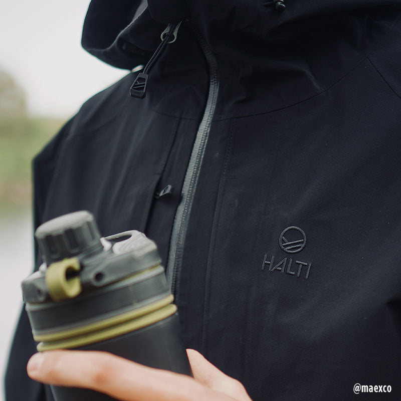 A person wears the Halti Kaarna Mens DX 3L Shell Jacket and holds a black bottle with yellow accents.