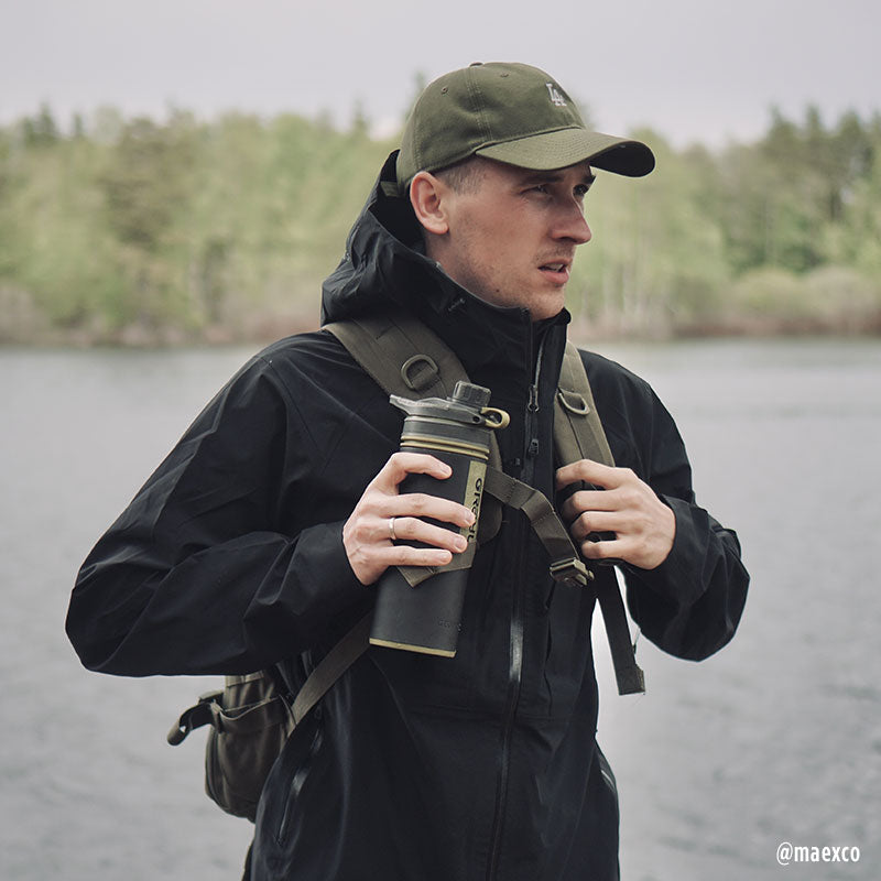 A man by a lake holds a drink bottle, wearing the Halti Kaarna Mens DX 3L Shell Jacket, ready for adventure.