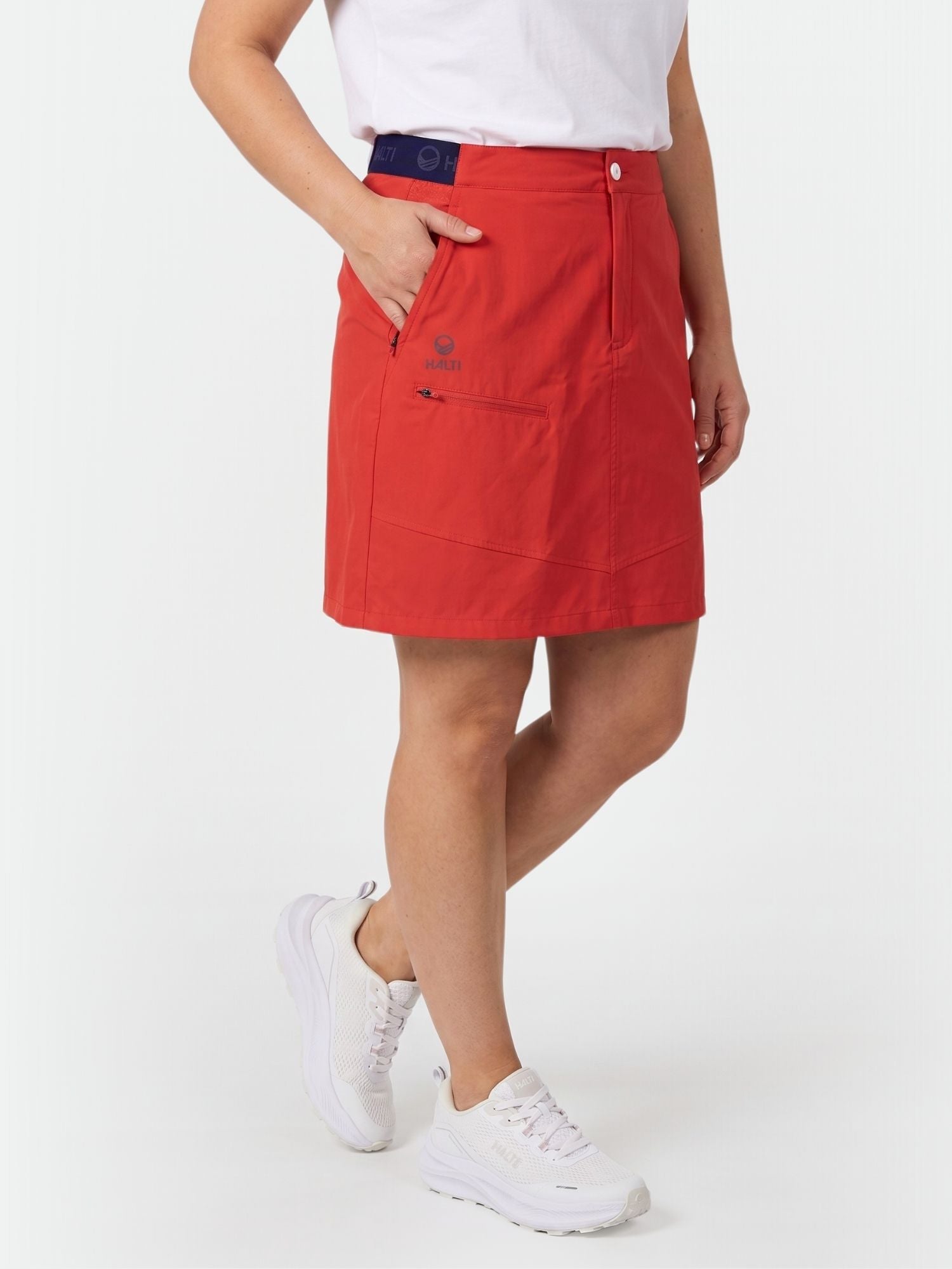 A person in a Halti Pallas Women's Lite X-stretch Skort, white T-shirt, and sneakers stands with one hand in their pocket.