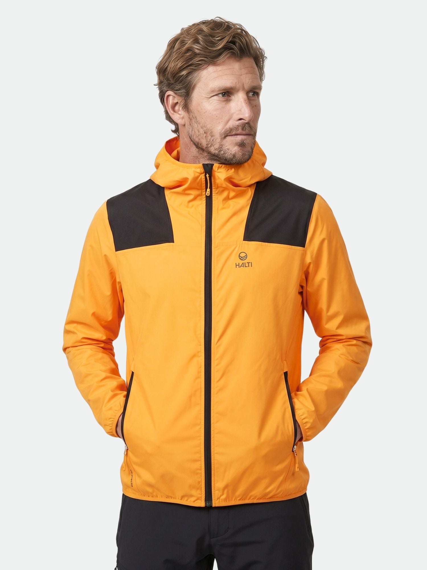 Man wearing Halti Pallas Men's Lite X-stretch jacket with black details and hood, hands in pockets, against plain background.