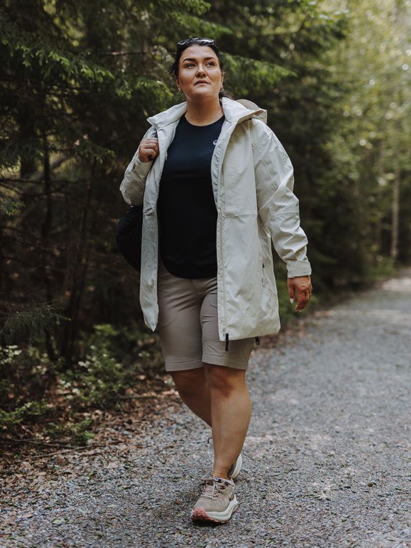 A woman wearing Halti Reissu Plus Womens Stretch Shorts and sneakers walks on a gravel path in the woods.