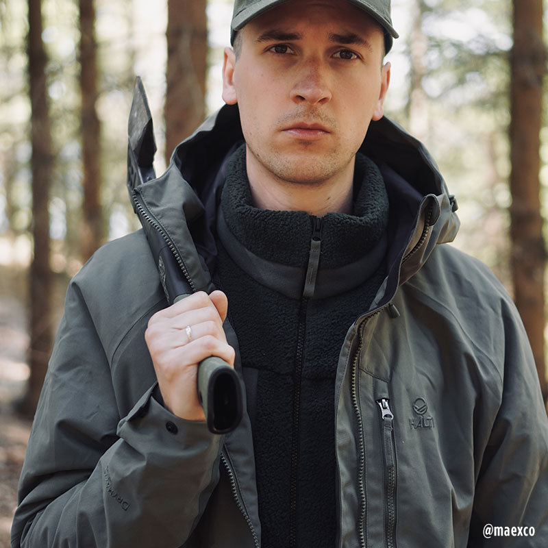 A man wearing the Halti Hiker Mens DrymaxX Pro Jacket stands in a forest, holding a strap and facing the camera.