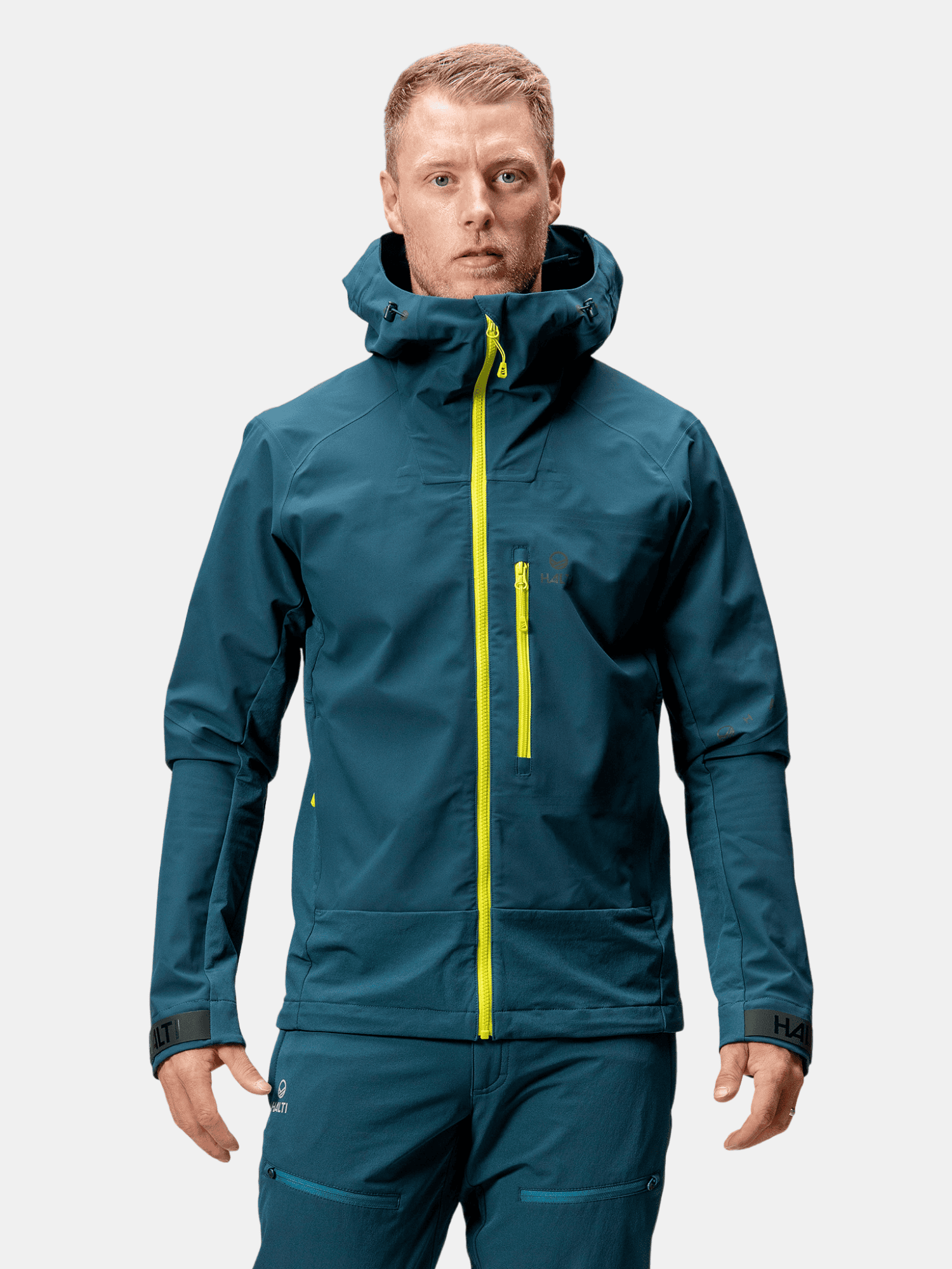 A man wears the Halti Pallas III Men's Warm Hybrid Jacket in teal with matching pants and yellow zippers, outdoors.