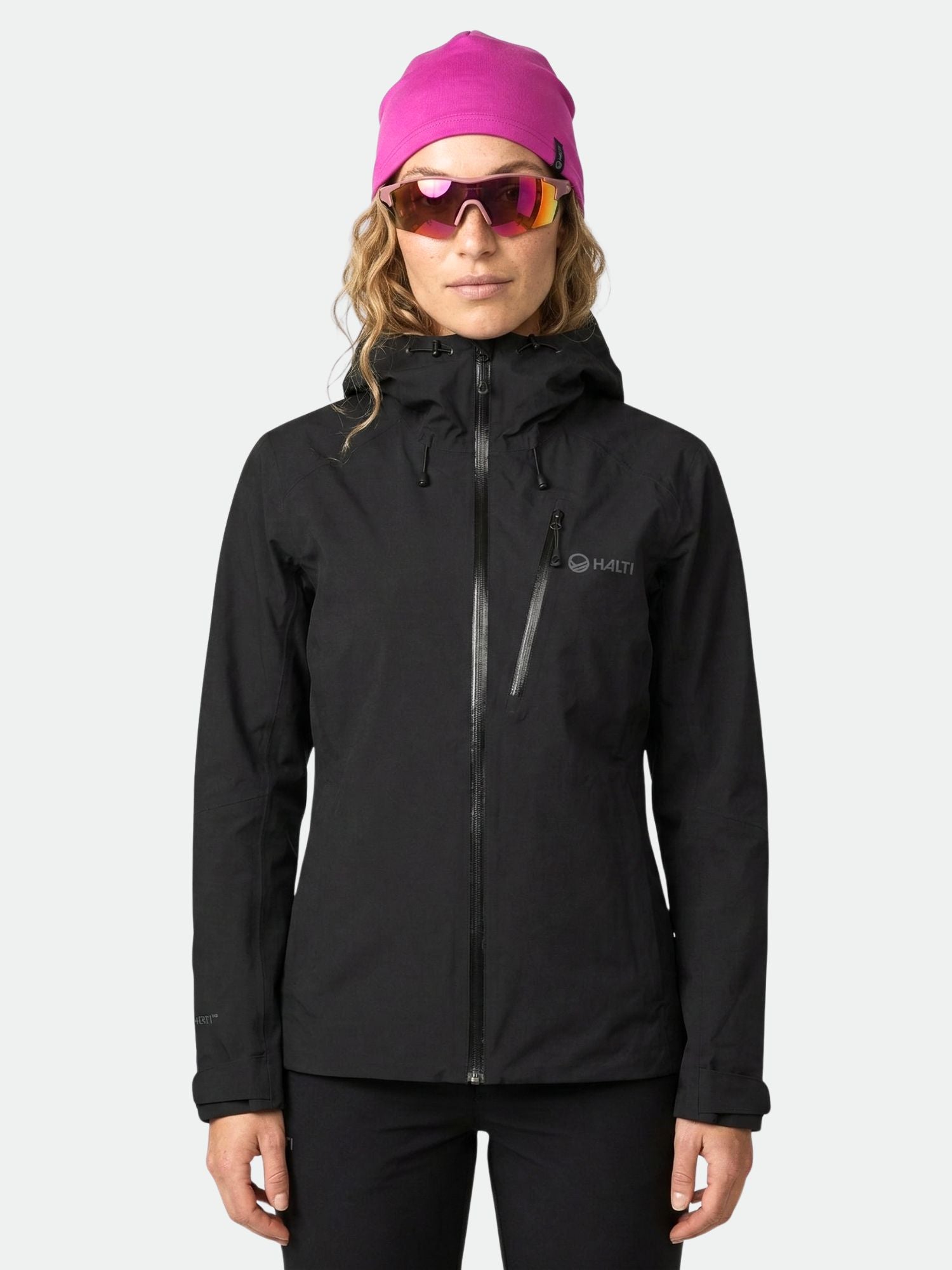 A woman in a Halti Biegga Women's DrymaxX Shell Jacket, pink beanie, and sunglasses stands against a light background.