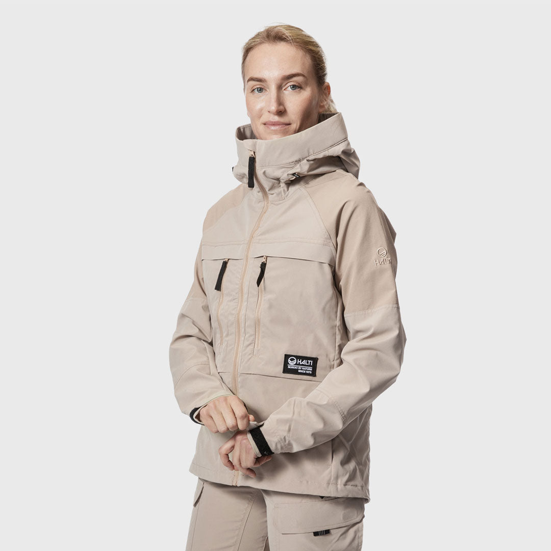 A woman wears the Halti Hiker Womens Lite Outdoor Jacket in beige, featuring a high collar and multiple pockets.
