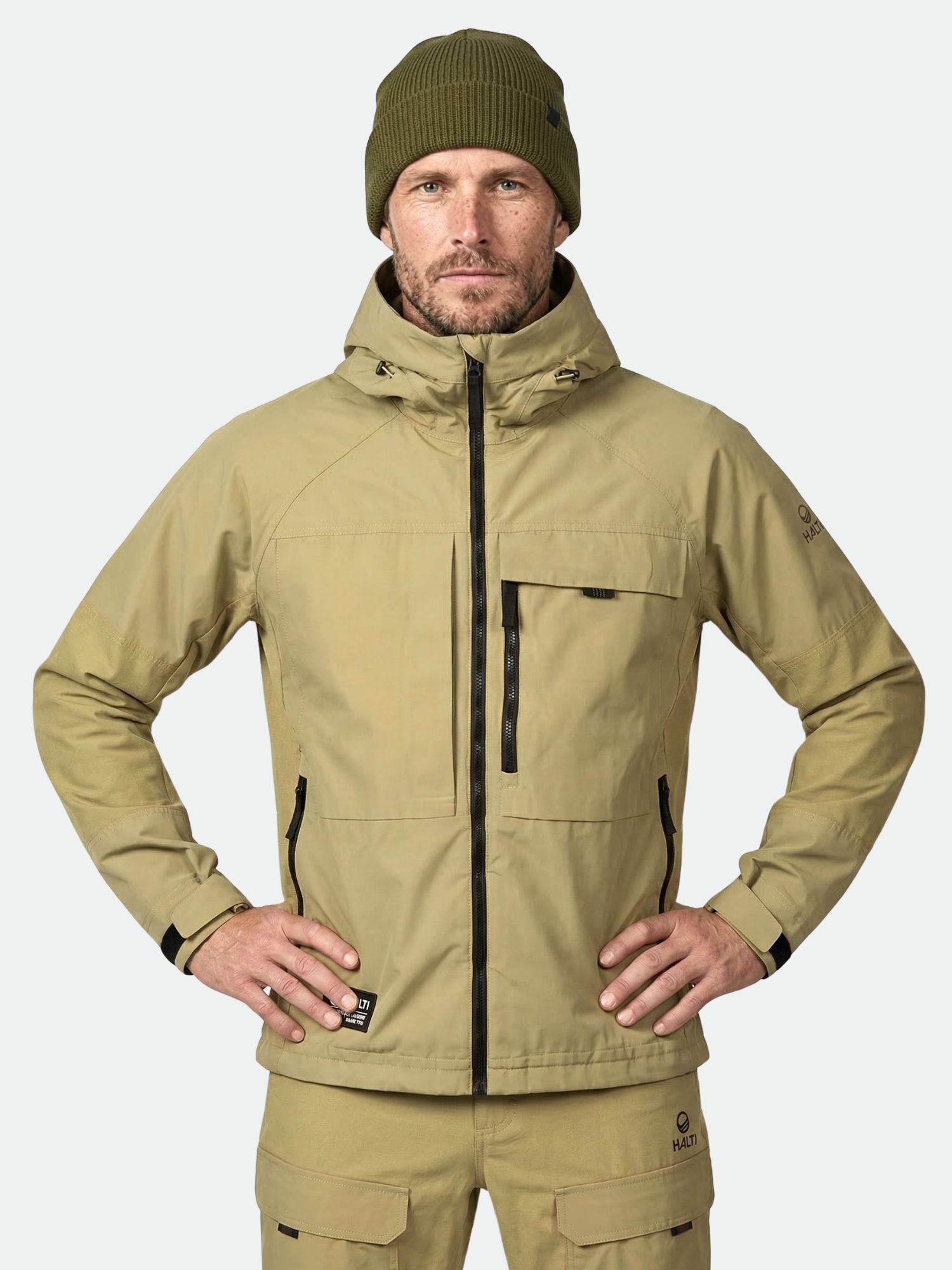 Man in Halti Hiker Men's Ventilated Jacket with matching pants and olive green beanie, hands on hips, plain background.