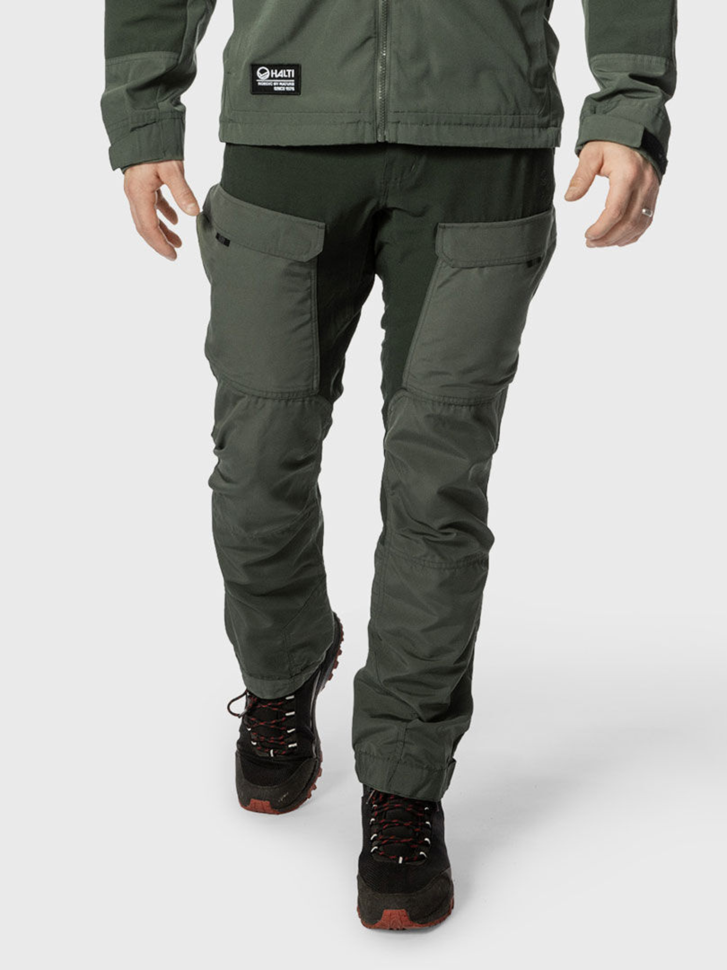 A person in Halti Hiker Men's Ventilated Pants and a matching jacket with black hiking shoes stands against a white background.