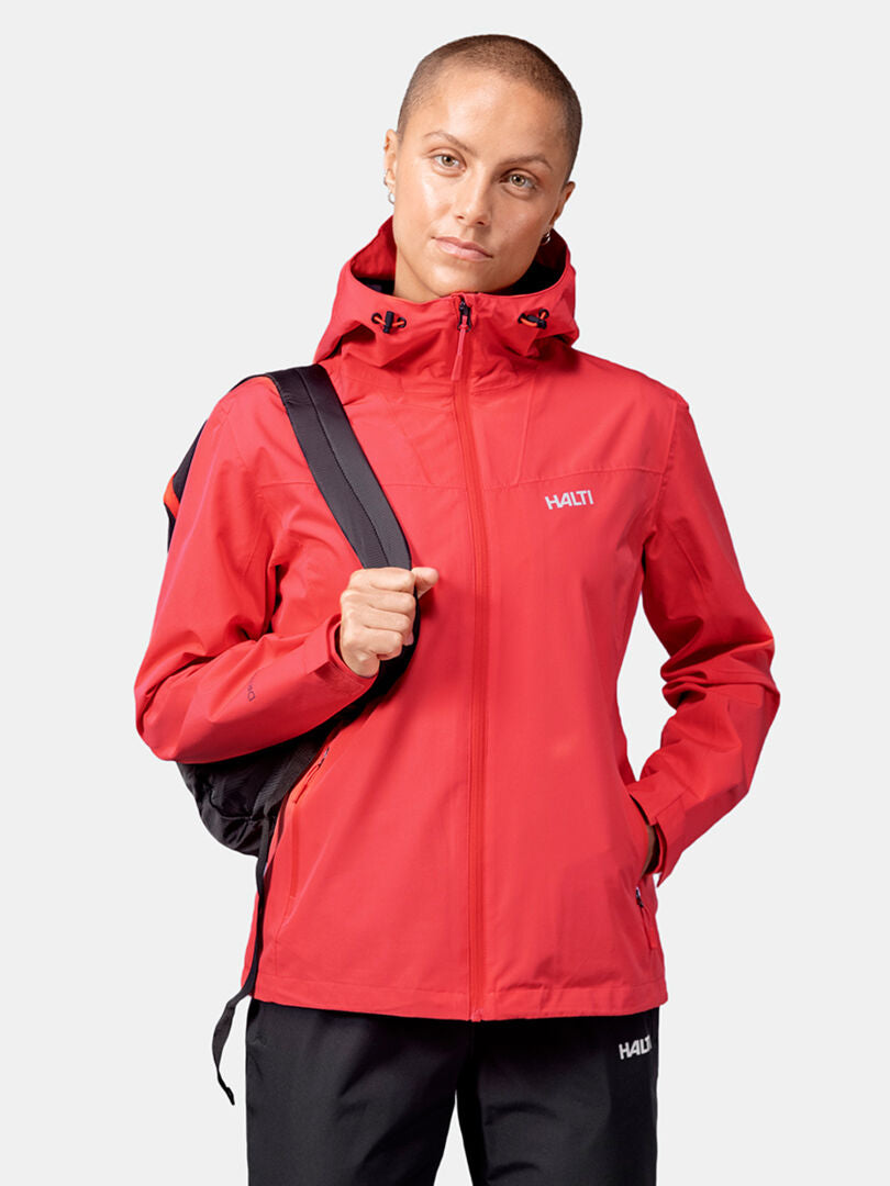Woman wearing a red Halti Forter DX Shell Jacket with hood and black pants, holding a backpack strap against a plain background.