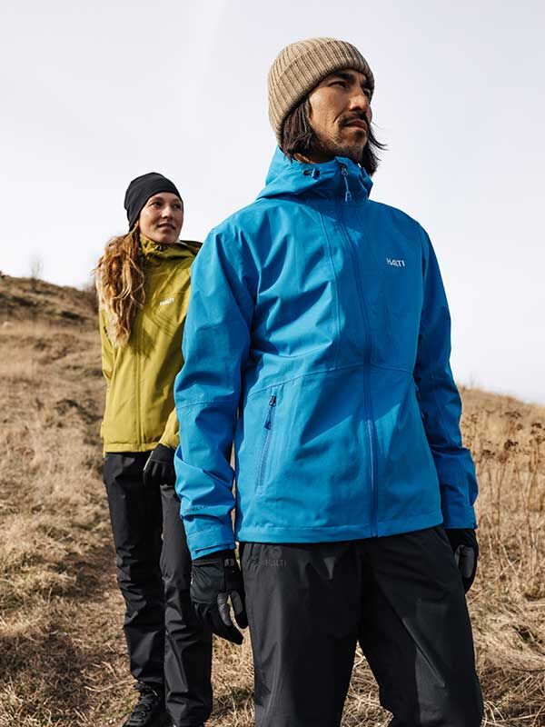 Two people in Halti Forter DX Shell jackets and beanies stand on a grassy hill, gazing into the cloudy distance.