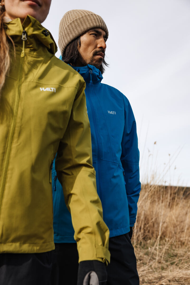 Two people in Halti Forter DX Shell Men's jackets—one yellow-green, one blue—stand in a grassy field under clouds.