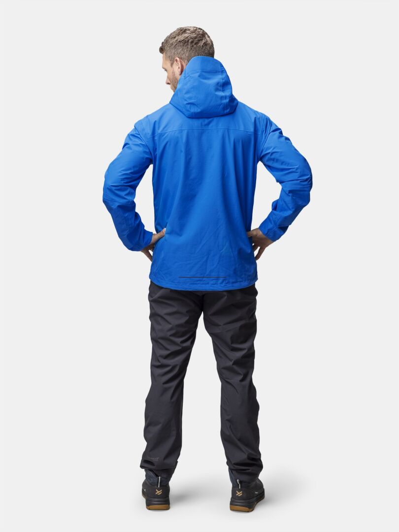 A man wears the Halti Forter DX Shell Jacket in blue and black pants, standing against a plain light background.
