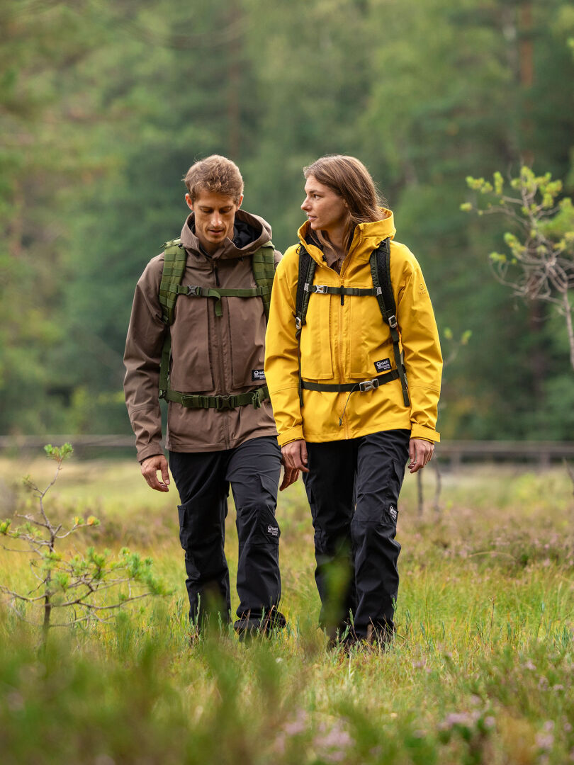 Two people wearing Halti Hiker II DrymaxX Shell Pants hike a grassy trail with backpacks amid green trees.