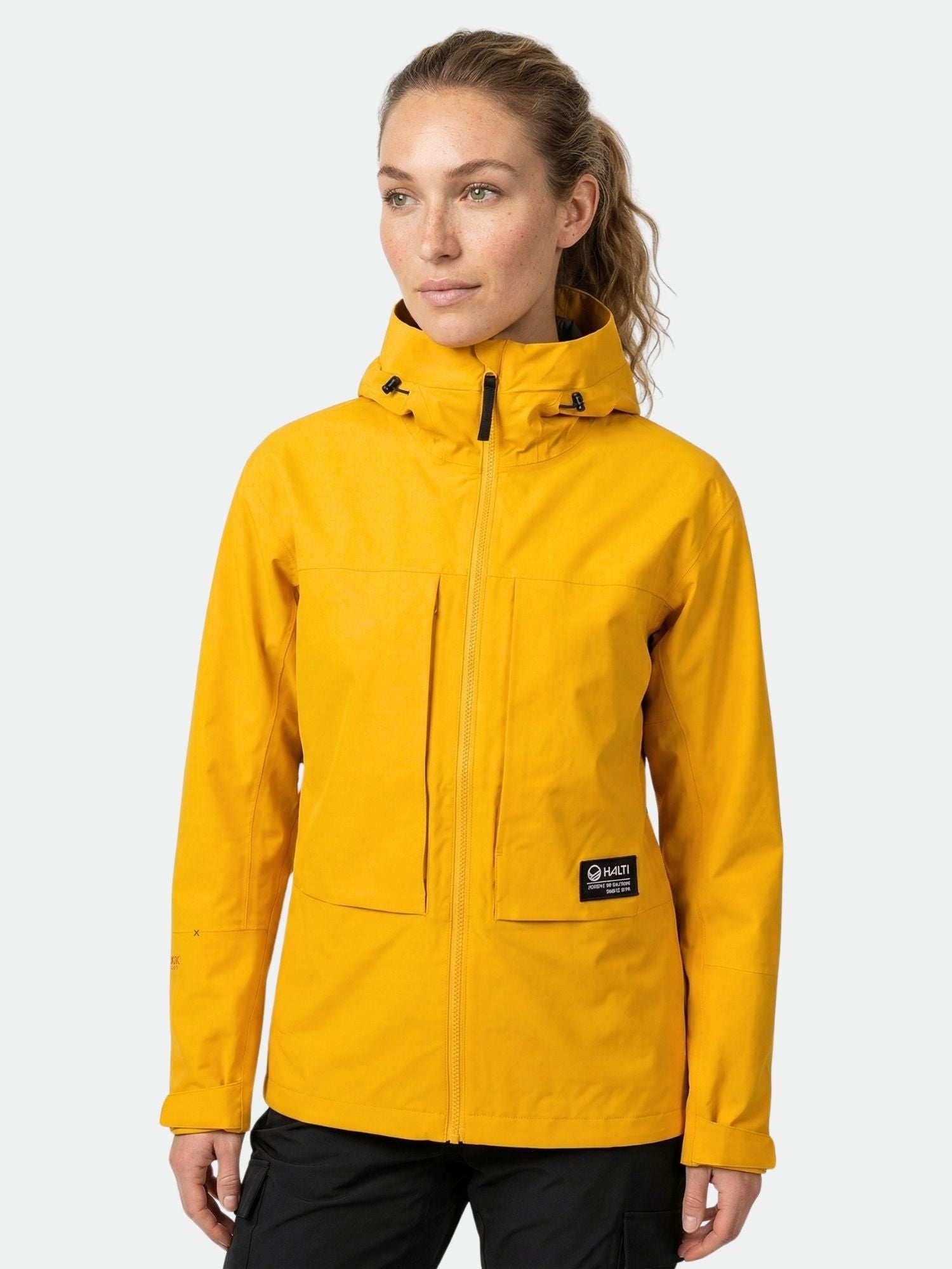 A woman wears a yellow Halti Hiker II DrymaxX Shell Jacket with black pants, standing against a plain background.