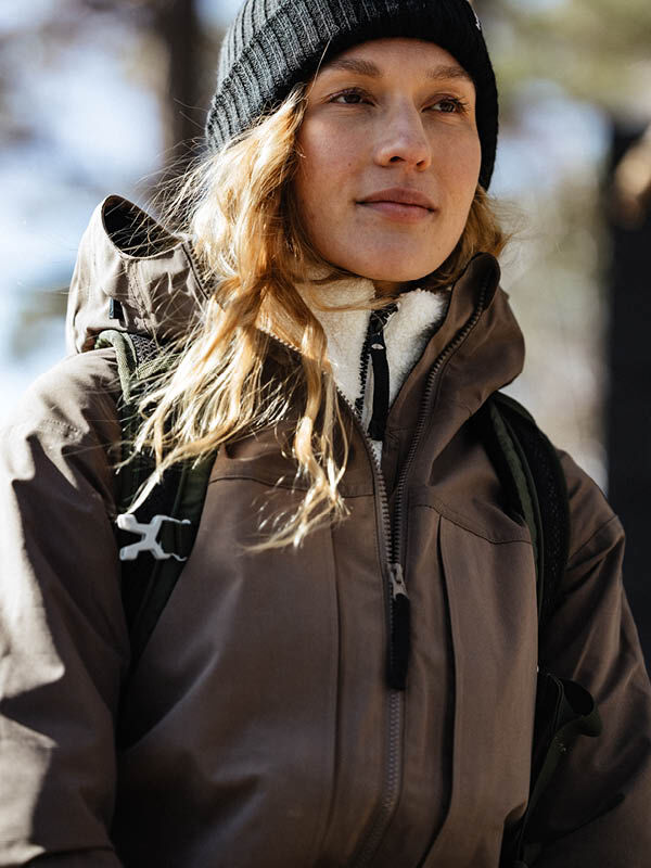 Wearing the Halti Runko Beanie and a brown jacket, a person stands outdoors with a backpack, gazing ahead.