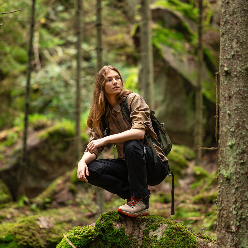 A person with long hair squats on a mossy rock in a forest, wearing Halti Hiker II DrymaxX Shell Pants for women.