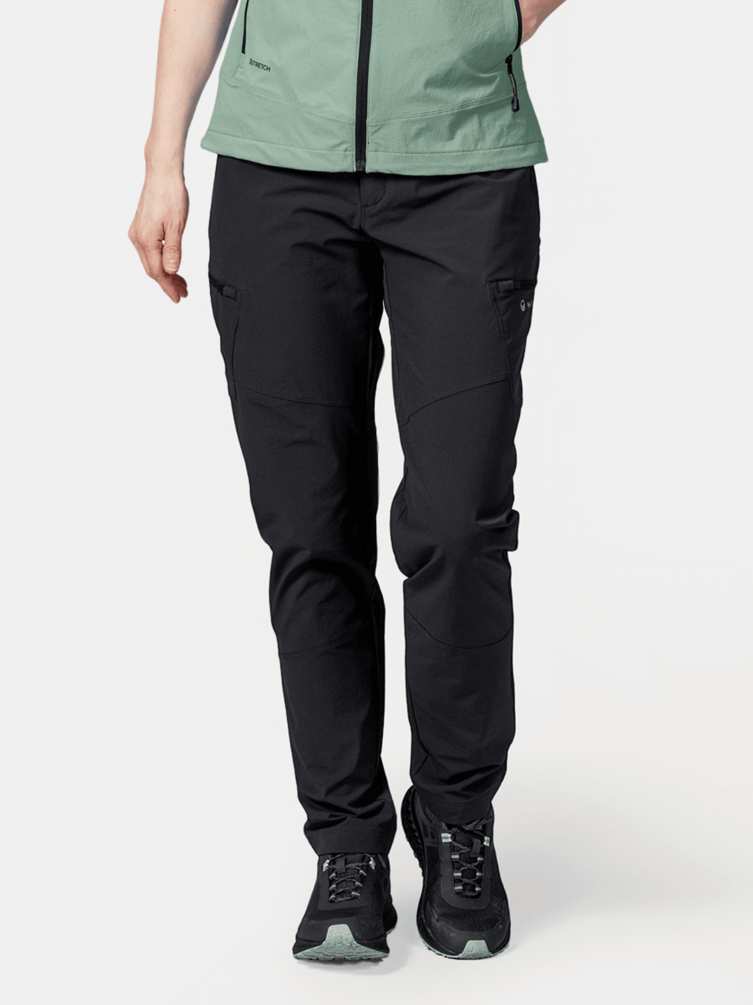 A person wears Halti Pallas Evo X-stretch Pants, black sneakers, and a green zip-up jacket against a white background.