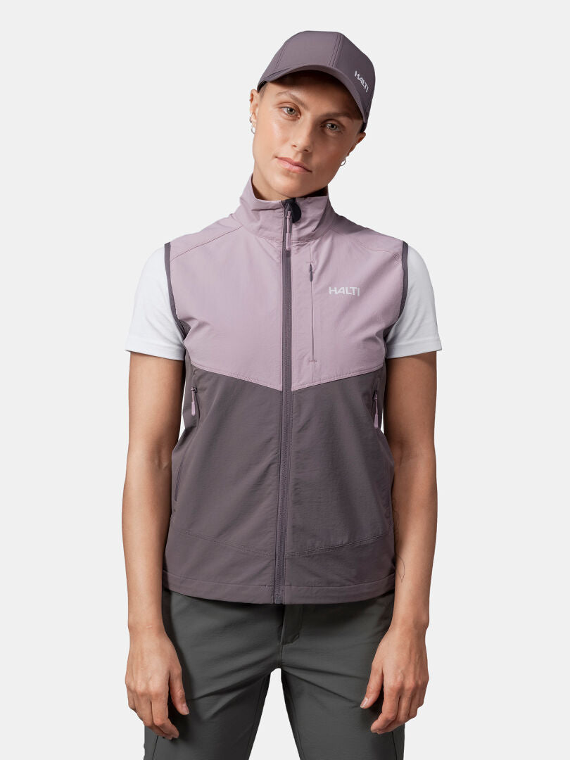Woman wearing Halti Pallas Evo X-stretch Vest in mauve/gray, white tee, gray pants, and matching cap on plain background.