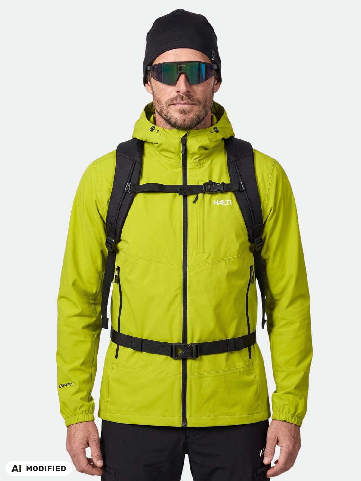 Man wearing Halti Pallas Evo X-stretch Jacket, black hat, sunglasses, and backpack on white background.